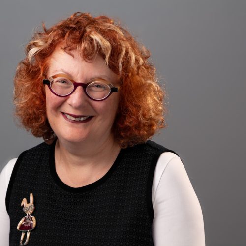 Portrait of Julie Abramson, who has curly red hair. She is wearing glasses and a black sleeveless top over a white long-sleeve shirt, with a bunny pin.